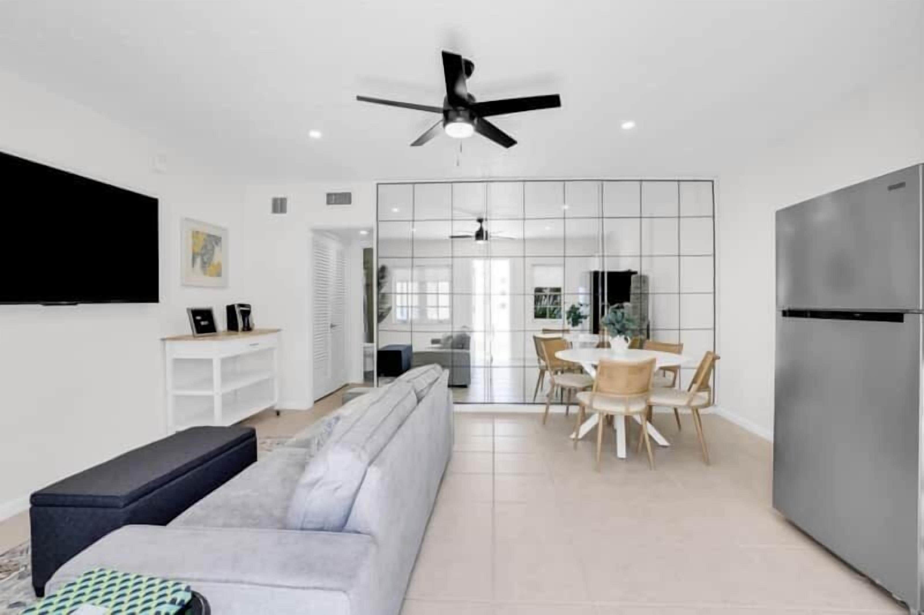 A modern living room with a grey sofa, a black bench, a dining area with a white table and chairs, and a large flat-screen TV mounted on the wall.