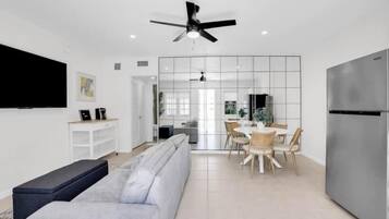 A modern living room with a grey sofa, a black bench, a dining area with a white table and chairs, and a large flat-screen TV mounted on the wall.