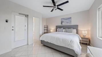 Un dormitorio con cama, ventilador de techo, mesita de noche con lámpara y puerta con mosquitera.