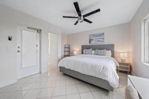Un dormitorio con cama, ventilador de techo, mesita de noche con lámpara y puerta con mosquitera.