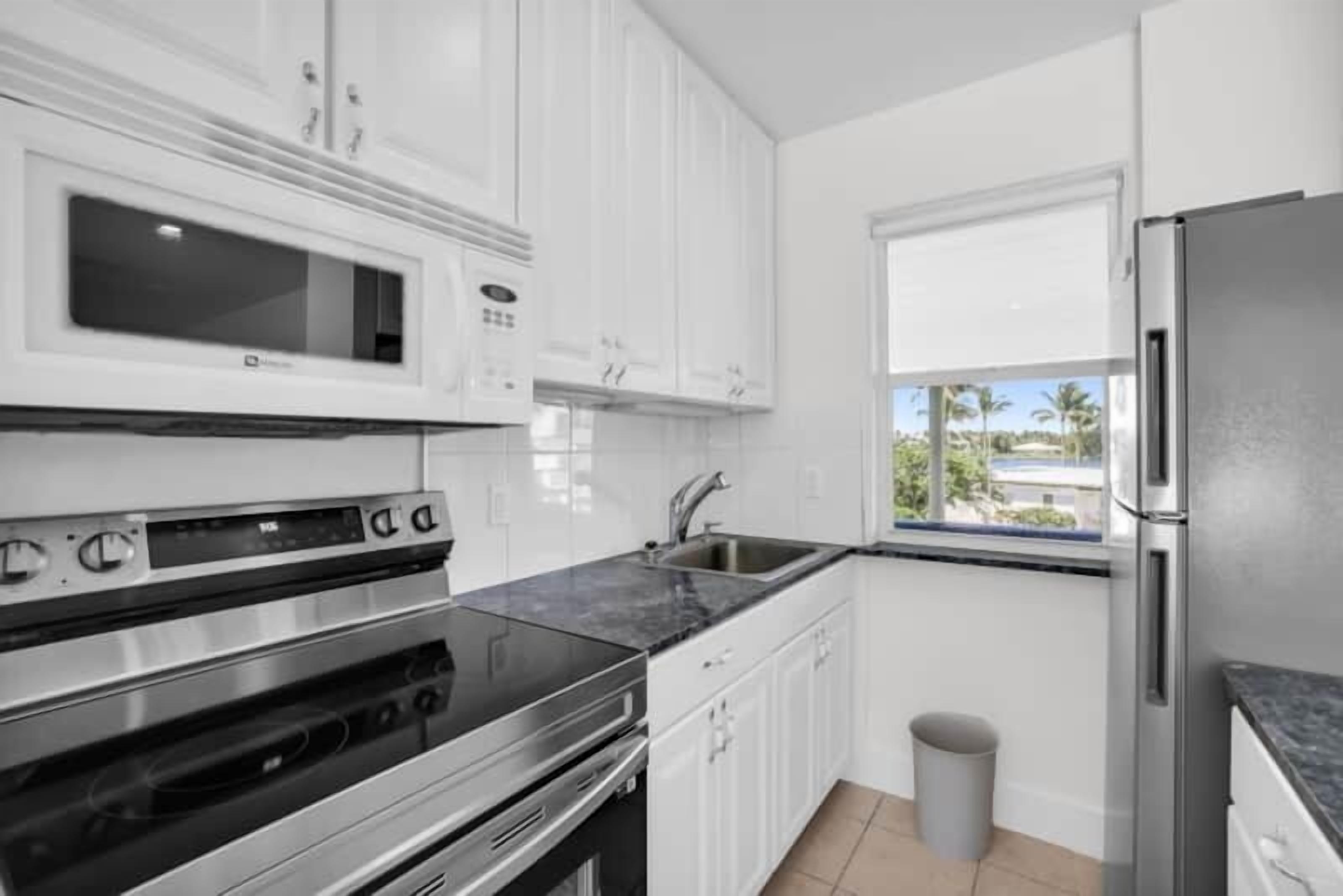 A modern kitchen with white cabinets, a stainless steel refrigerator, a built-in microwave, and a window with a view of palm trees.
