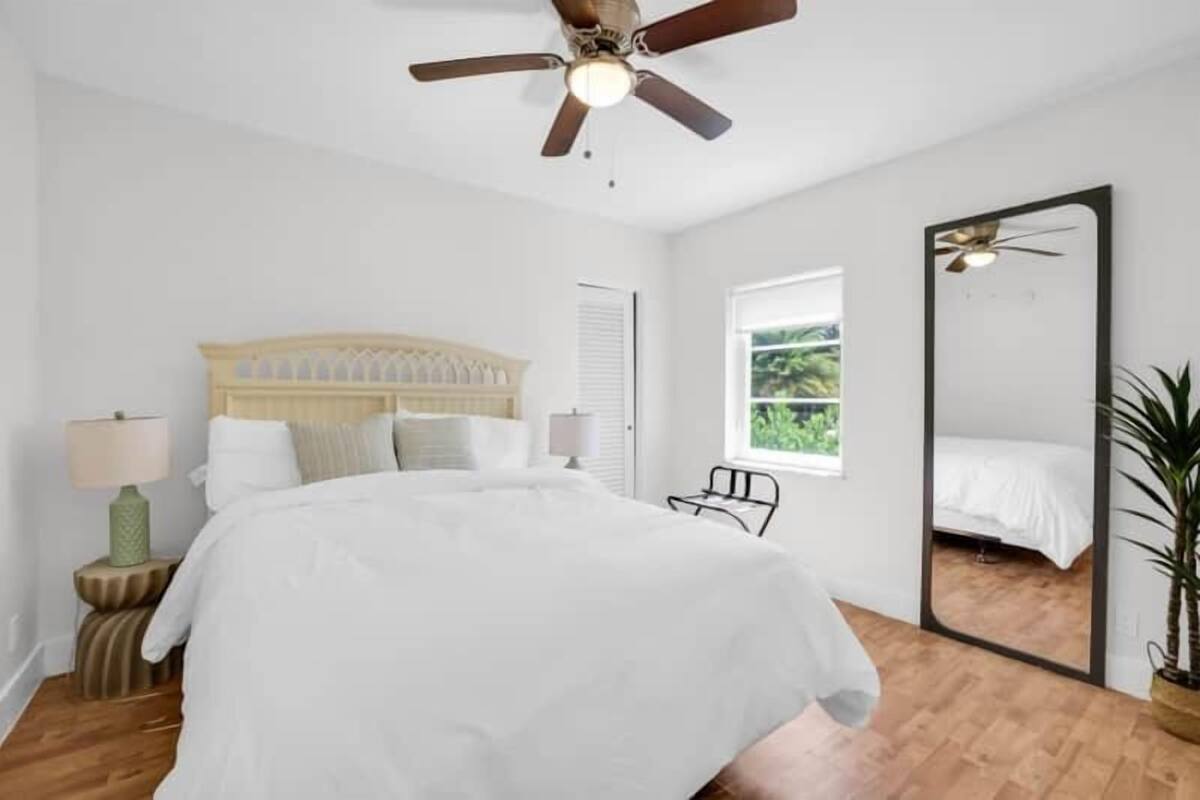Un dormitorio con una cama, ventilador de techo, ventana, espejo y planta.