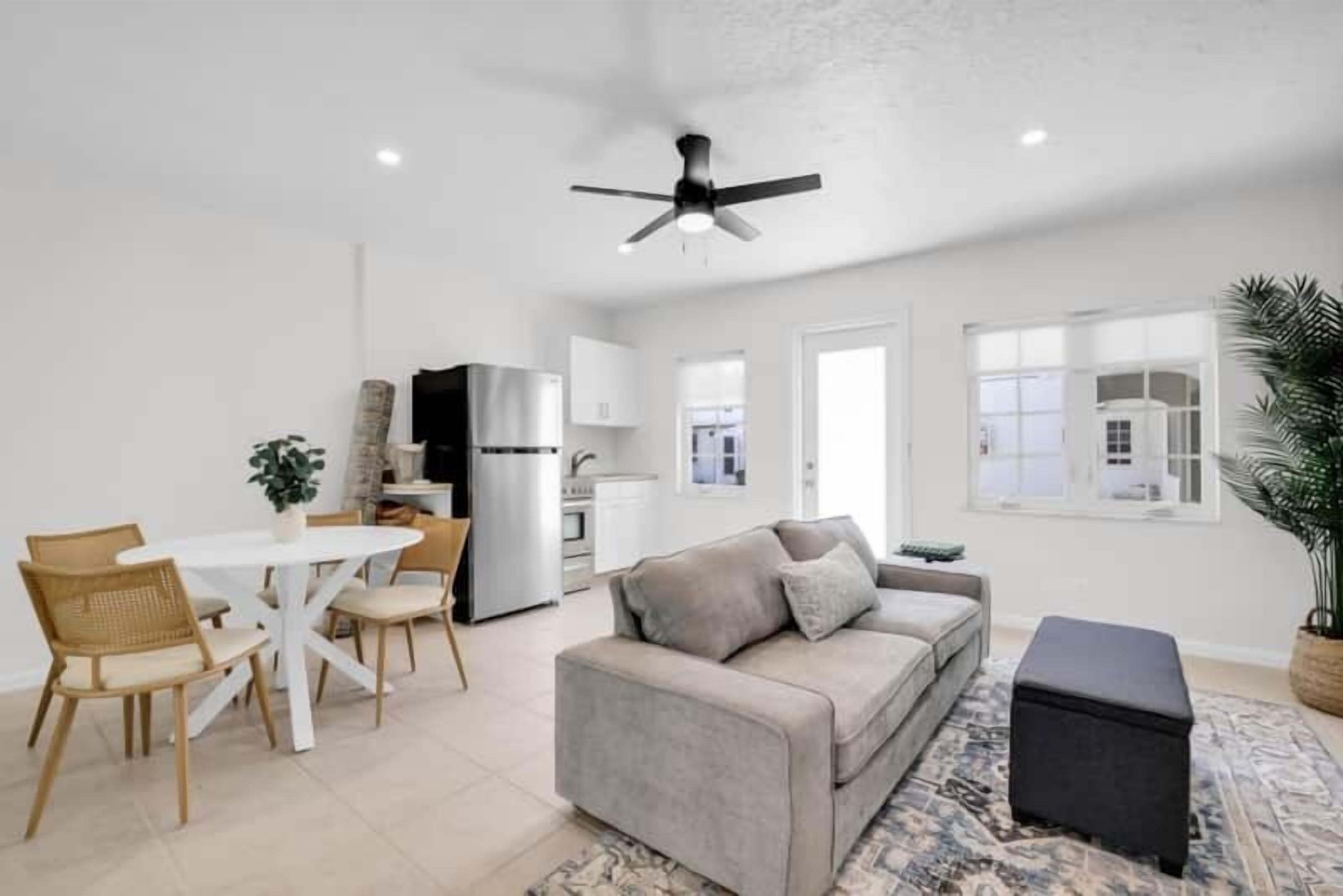 A modern living room with a grey sofa, a dining table with chairs, a ceiling fan, and a kitchen in the background.