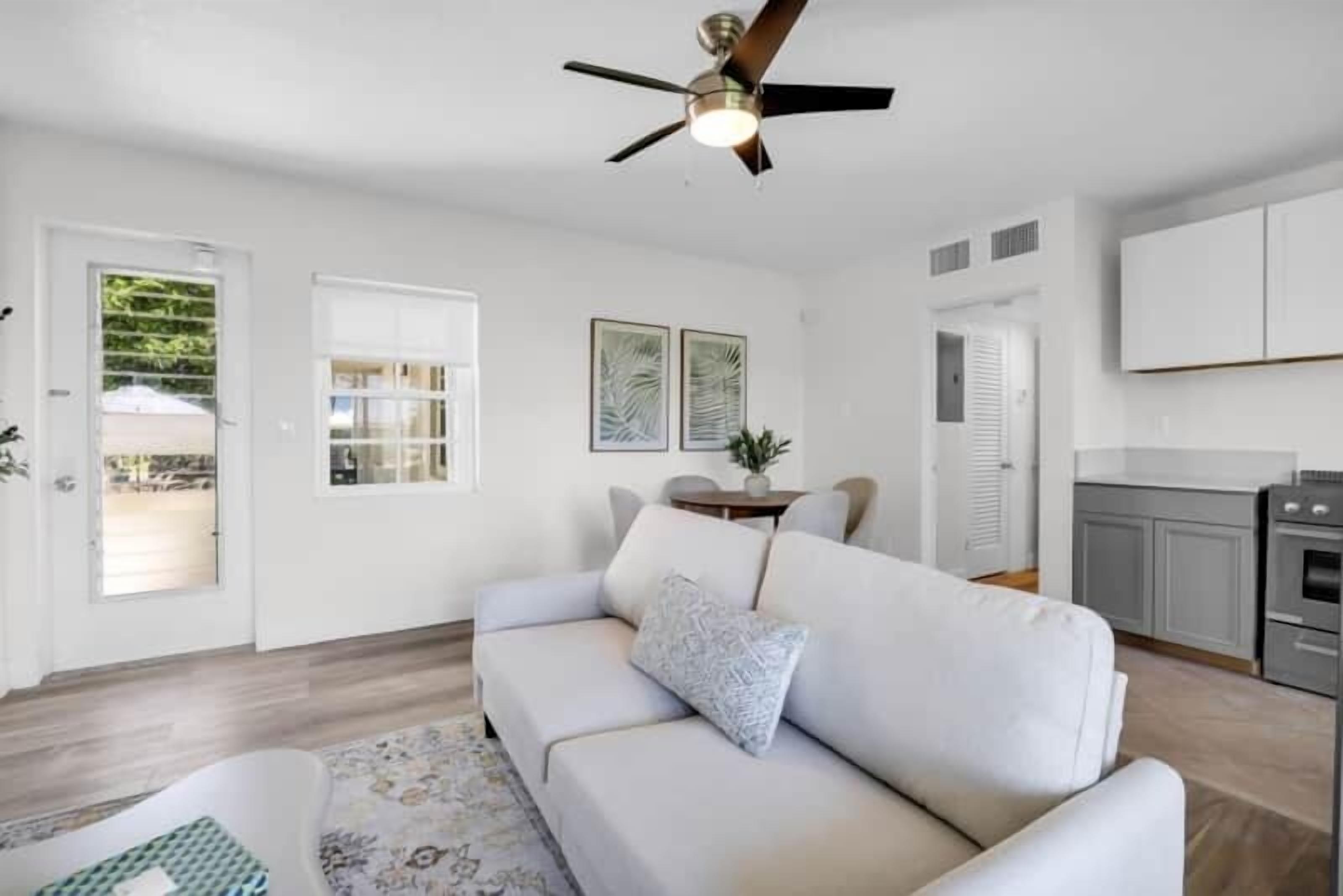 A modern living room with a white sofa, a ceiling fan, and a kitchen area in the background.