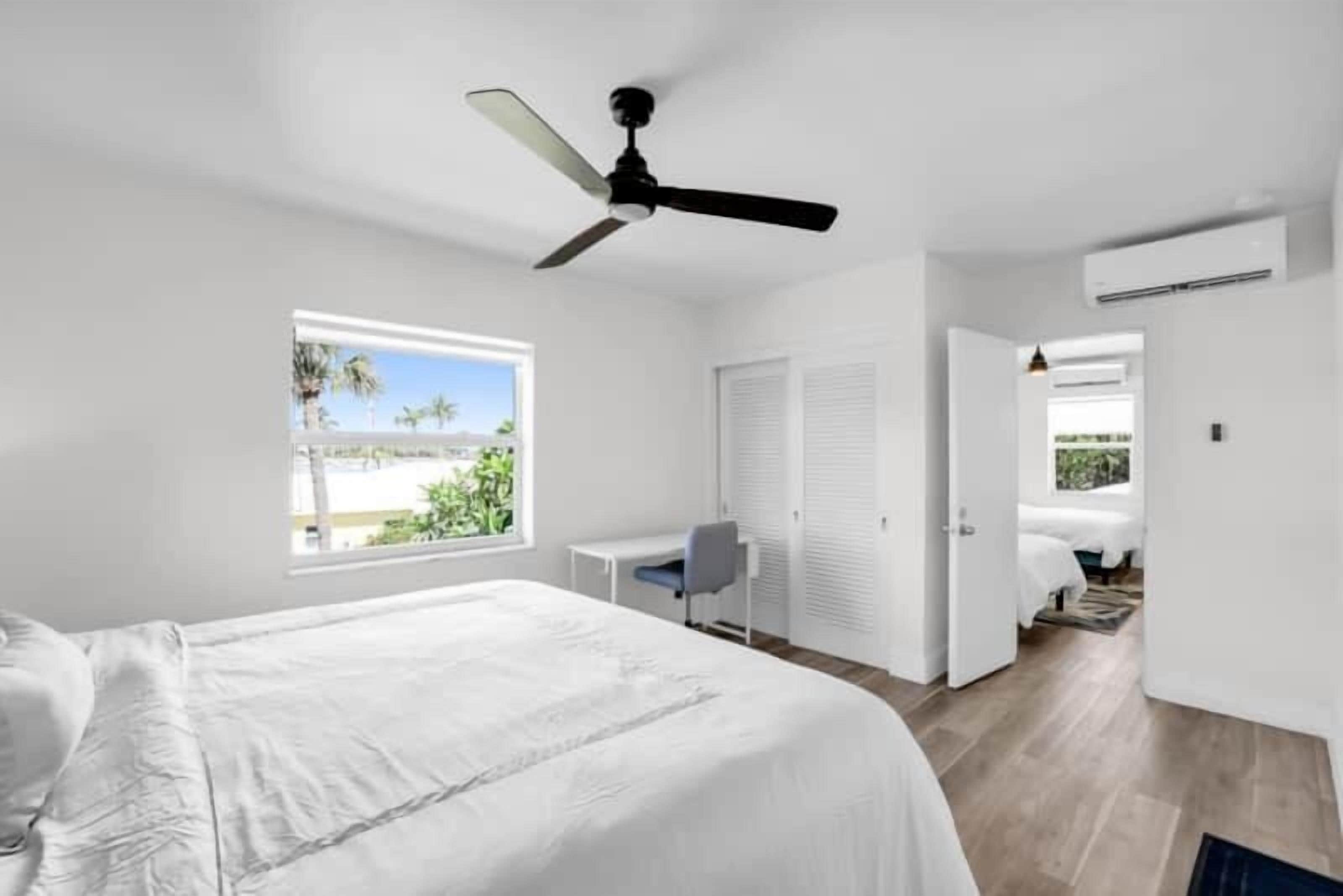 A bedroom with a bed, a desk, a chair, a ceiling fan, and a window with a view of palm trees.