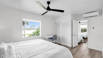 A bedroom with a bed, a desk, a chair, a ceiling fan, and a window with a view of palm trees.