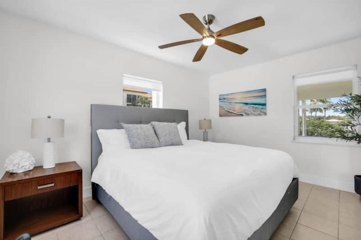 Un dormitorio con cama, mesita de noche, ventilador de techo, ventana y un cuadro en la pared.
