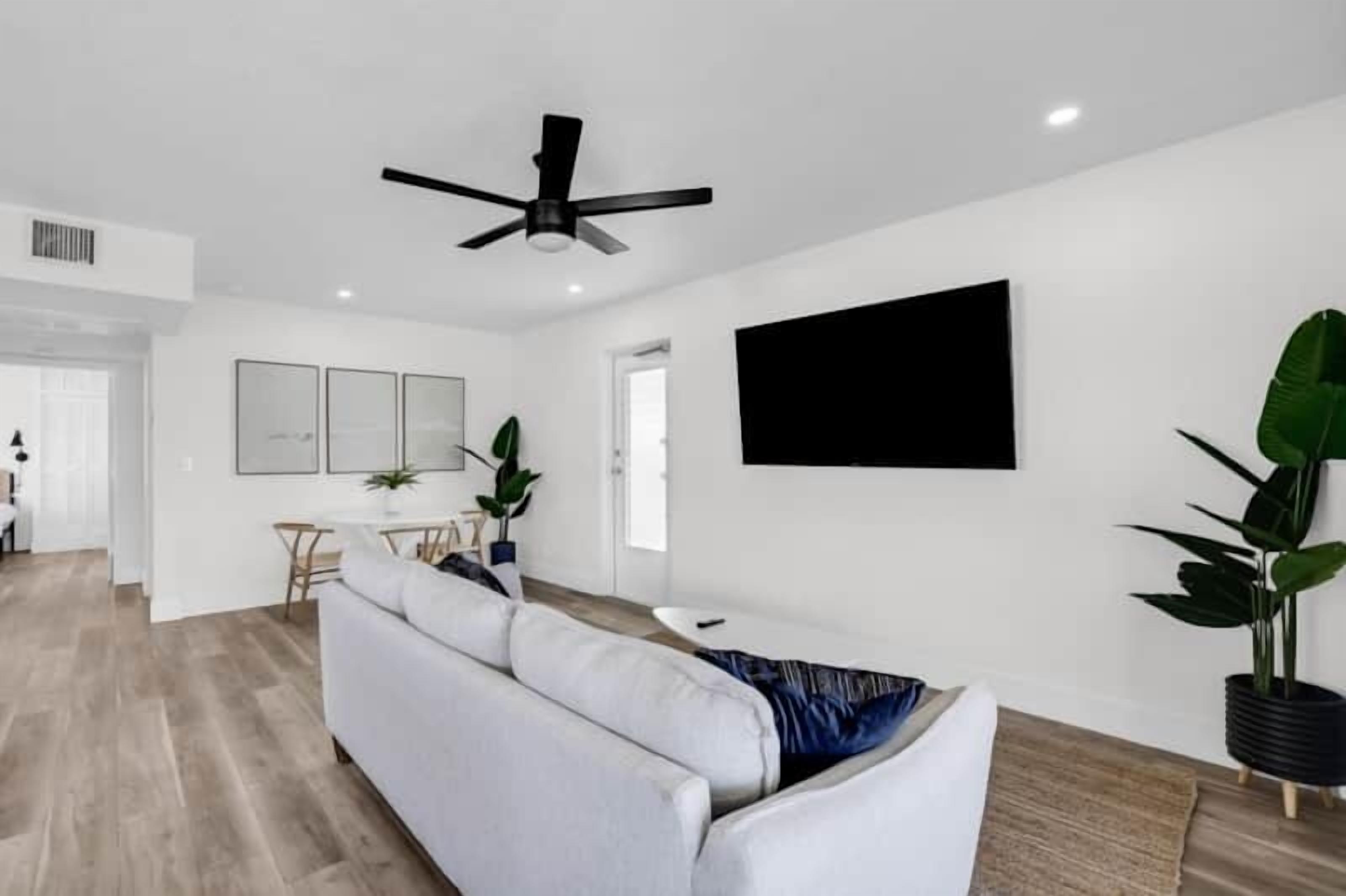A modern living room with a white sofa, a ceiling fan, a flat-screen TV, and a dining area with a table and chairs.