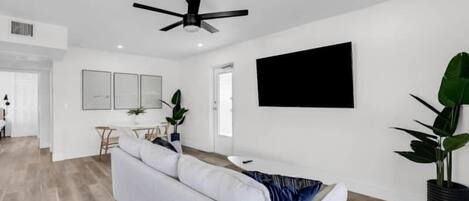 A modern living room with a white sofa, a ceiling fan, a flat-screen TV, and a dining area with a table and chairs.