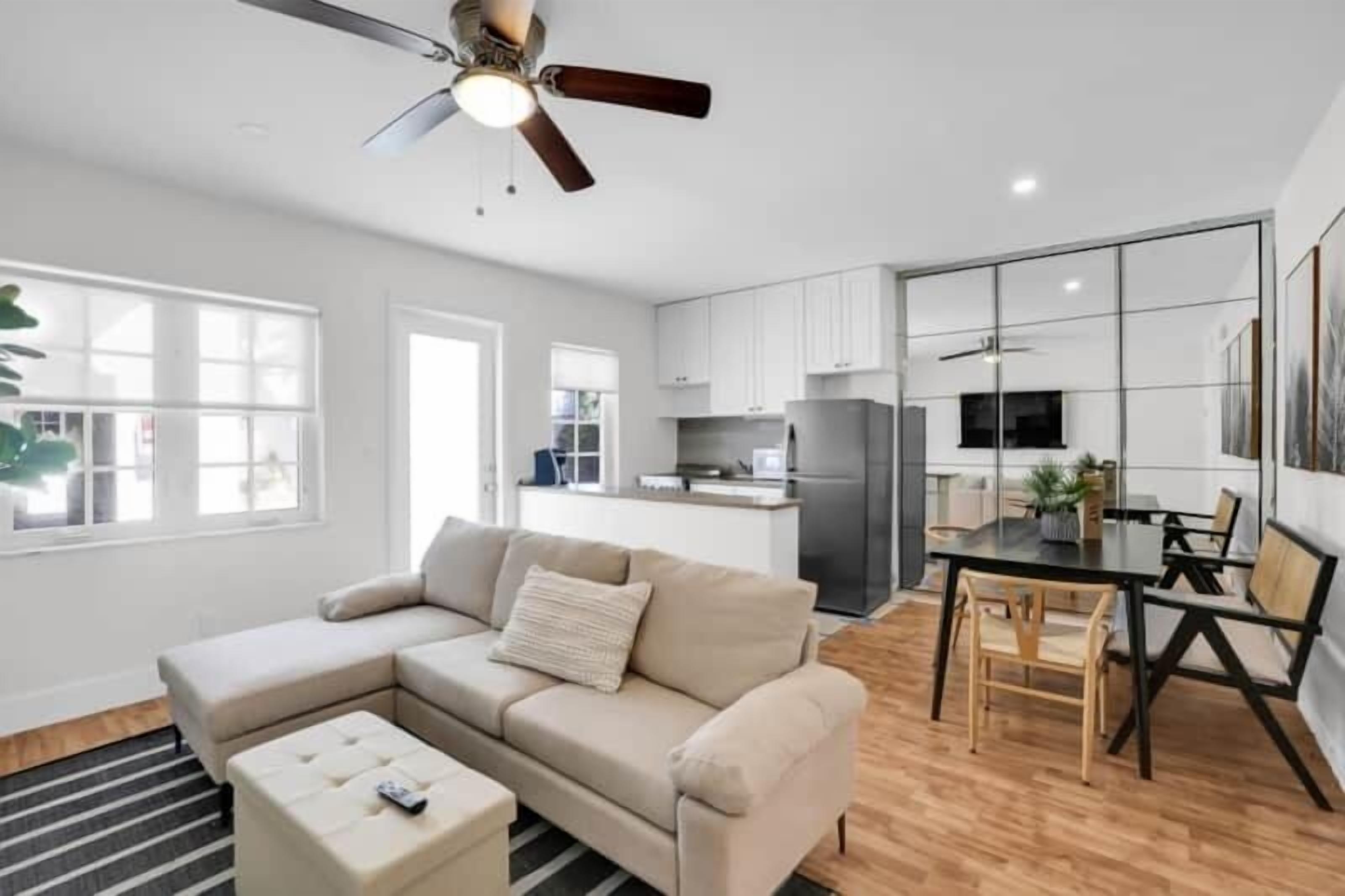 A modern living room with a sectional sofa, a dining table with chairs, a ceiling fan, and a kitchen area with cabinets and appliances.