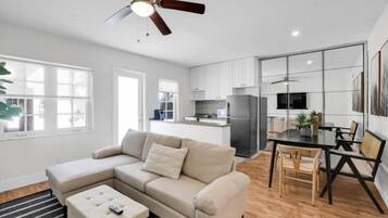 A modern living room with a sectional sofa, a dining table with chairs, a ceiling fan, and a kitchen area with cabinets and appliances.