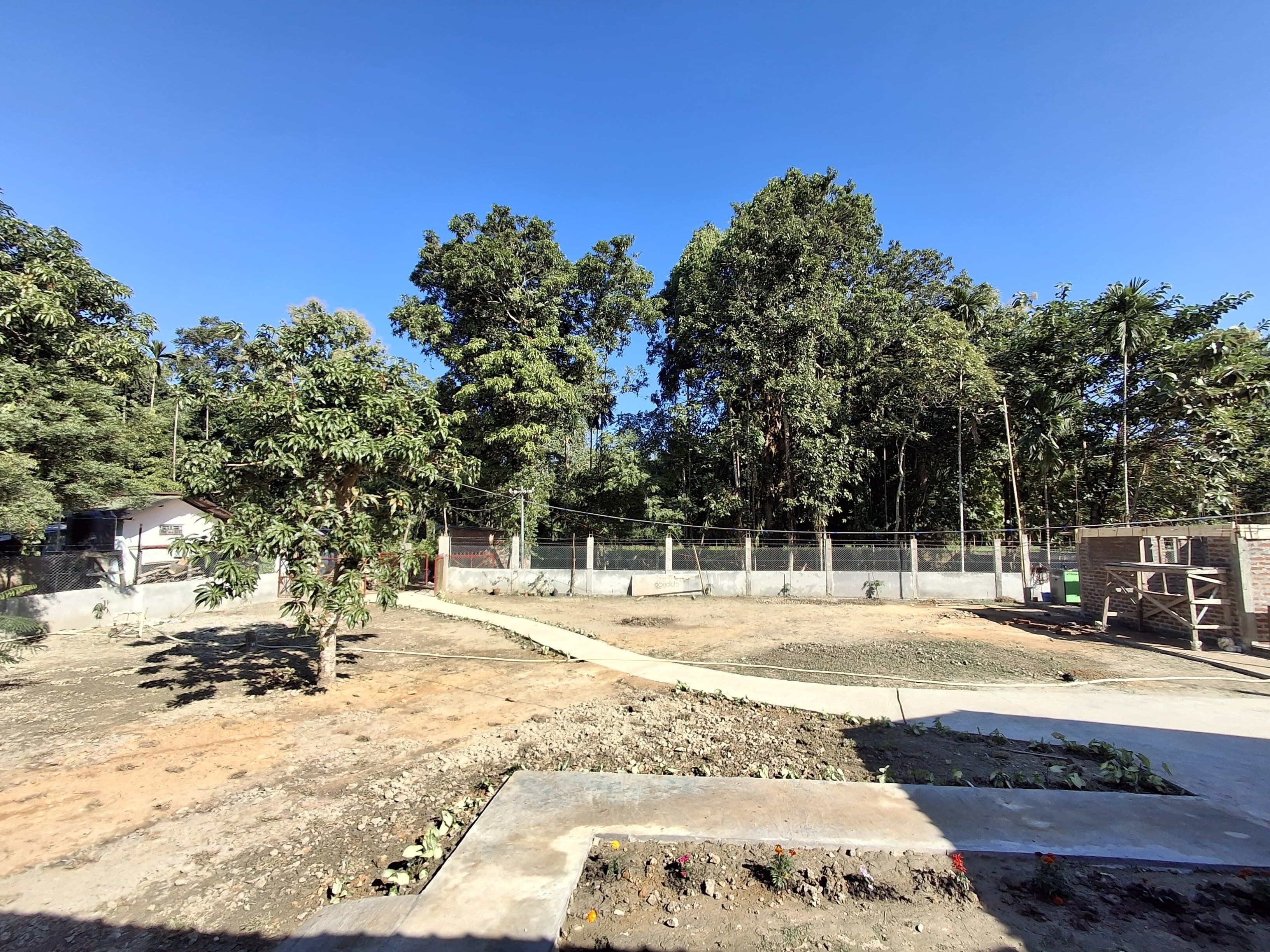 An outdoor area with a dirt ground, a concrete pathway, and a fenced enclosure with trees.