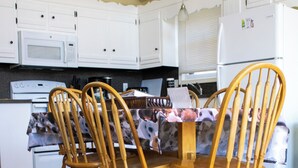 Een eetgedeelte met een houten tafel en stoelen, met op de achtergrond een keuken voorzien van witte kasten en apparatuur.
