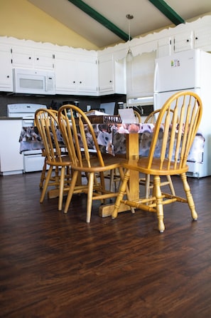 Un comedor con una mesa y sillas de madera, y una cocina al fondo con armarios y electrodomésticos blancos.