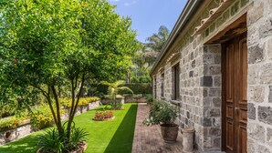 Un bâtiment en pierre avec une porte en bois, un chemin pavé et un jardin bien entretenu, agrémenté de diverses plantes et d’une pelouse.