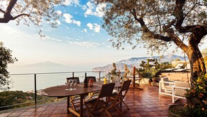 Uma área de refeições ao ar livre com mesa e cadeiras, com vista panorâmica para o mar e montanhas.