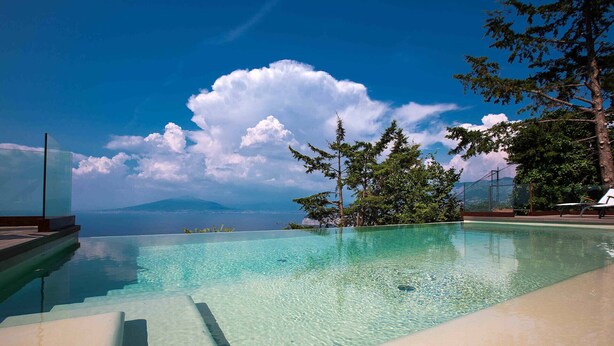 Uma piscina infinita e límpida com vista para o mar e montanhas, rodeada por árvores e um deck de madeira.