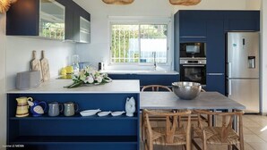 A modern kitchen with dark blue cabinets, a white countertop, and a large window. There are two hanging wicker lamps, a stainless steel refrigerator, and a dining area with a wooden table and chairs. - Sunset view, beach walk, heated pool (SAINT MARTIN)
