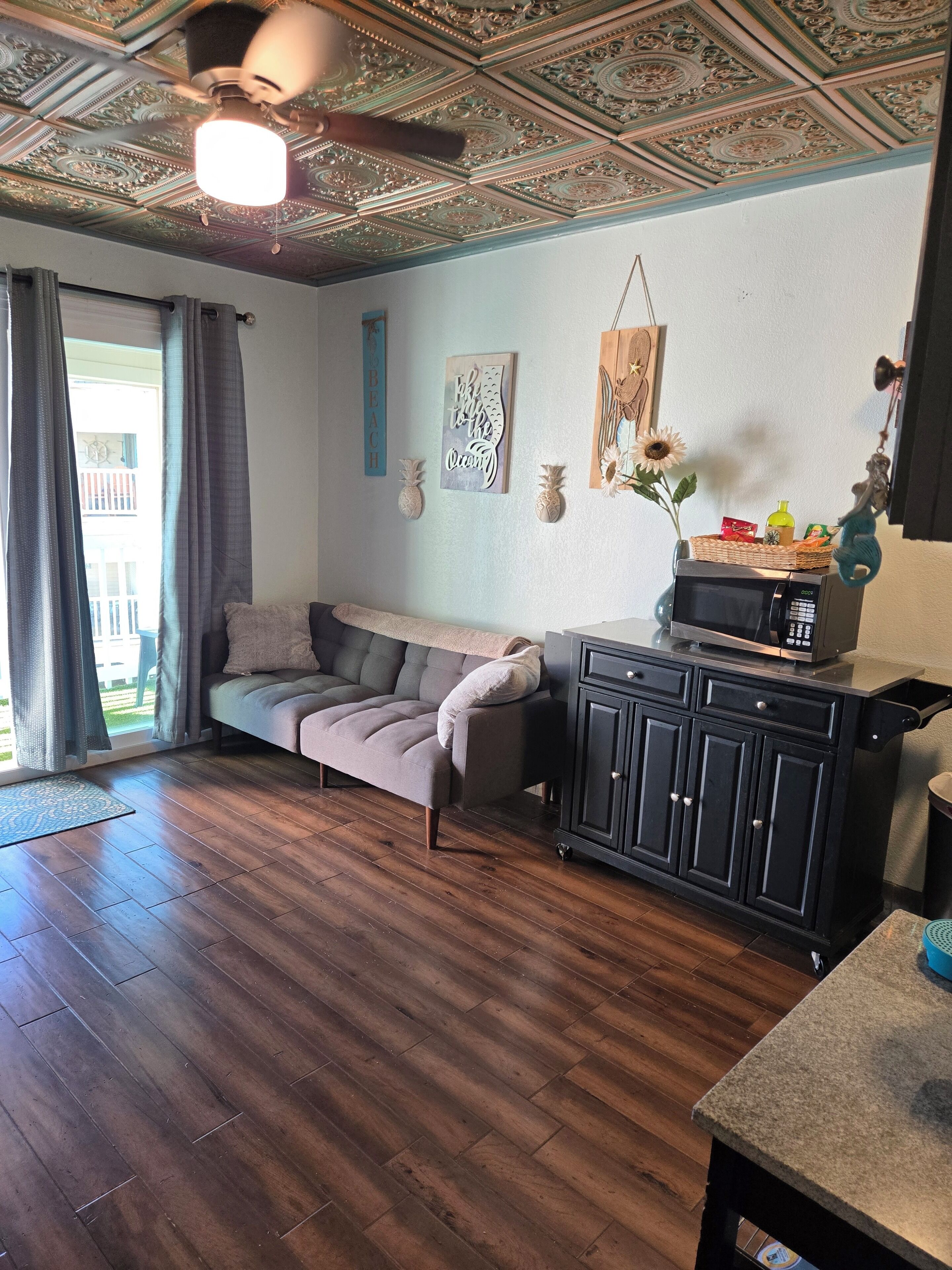 Una sala de estar con un sofá, piso de madera, un ventilador de techo y una zona de cocina con armarios y un microondas.