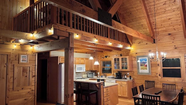 A cozy cabin interior with a wooden dining area, kitchen, and a staircase leading to an upper level. - Charming 3-bedroom + loft log cabin at Lake Bomoseen in Castleton, VT. Sleeps 12 (Fair Haven)