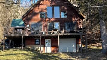 A two-story log cabin with a covered deck, a garage, and a large window overlooking a wooded area.