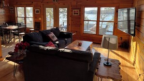 A cozy cabin interior with a wooden interior, large windows, a dining area, and a sofa. - Charming 3-bedroom + loft log cabin at Lake Bomoseen in Castleton, VT. Sleeps 12 (Fair Haven)