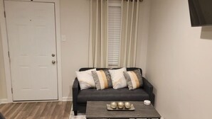 Ein Wohnzimmer mit einem dunkelgrauen Sofa, drei Kissen, einem Holztisch mit Dekoration, einer weißen Tür und einem Fenster mit weißen Vorhängen.
