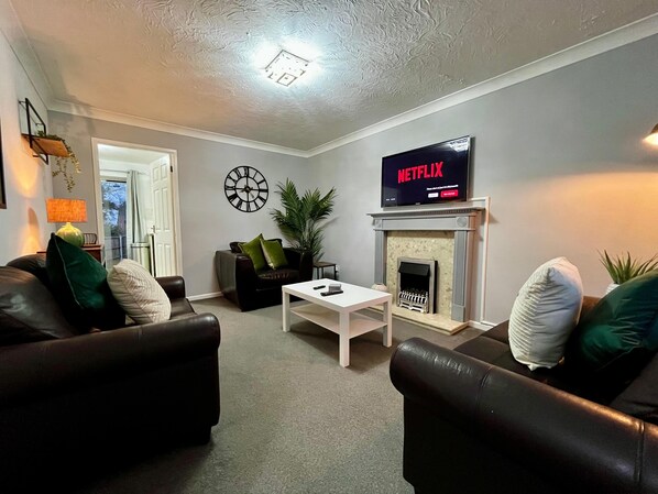 A living room with a fireplace, a television displaying 'NETFLIX', a clock, and a sofa. - 3 Bedroom Home in Telford (Newdale)