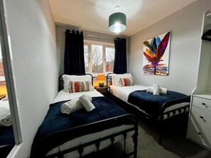 A bedroom with two single beds, a window with curtains, a bedside lamp, a colorful painting, and a small cabinet. - 3 Bedroom Home in Telford (Newdale)
