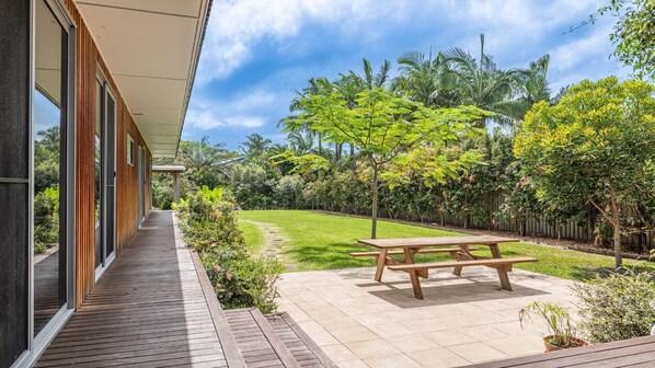 Una casa moderna con terraza de madera, mesa de picnic y un jardín bien cuidado con diversas plantas y árboles.