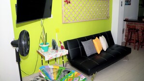 A living room with a black sofa, a small table with a floral tablecloth, a potted plant, a fan, and a TV mounted on a green wall. - Vacation apartment (Cartagena de Indias)