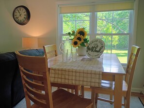 A dining area with a table set for four, a clock on the wall, and a window with a view of greenery.