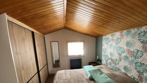Ein Schlafzimmer mit Holzboden, einem Bett mit gemusterten Betttextilien, einem Spiegel, einem Heizkörper und einem Fenster.