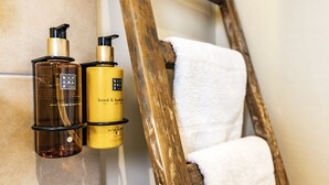 Dos botellas de jabón de manos y cuerpo sujetas a una pared, una silla de madera y una toalla blanca colgada en un perchero.