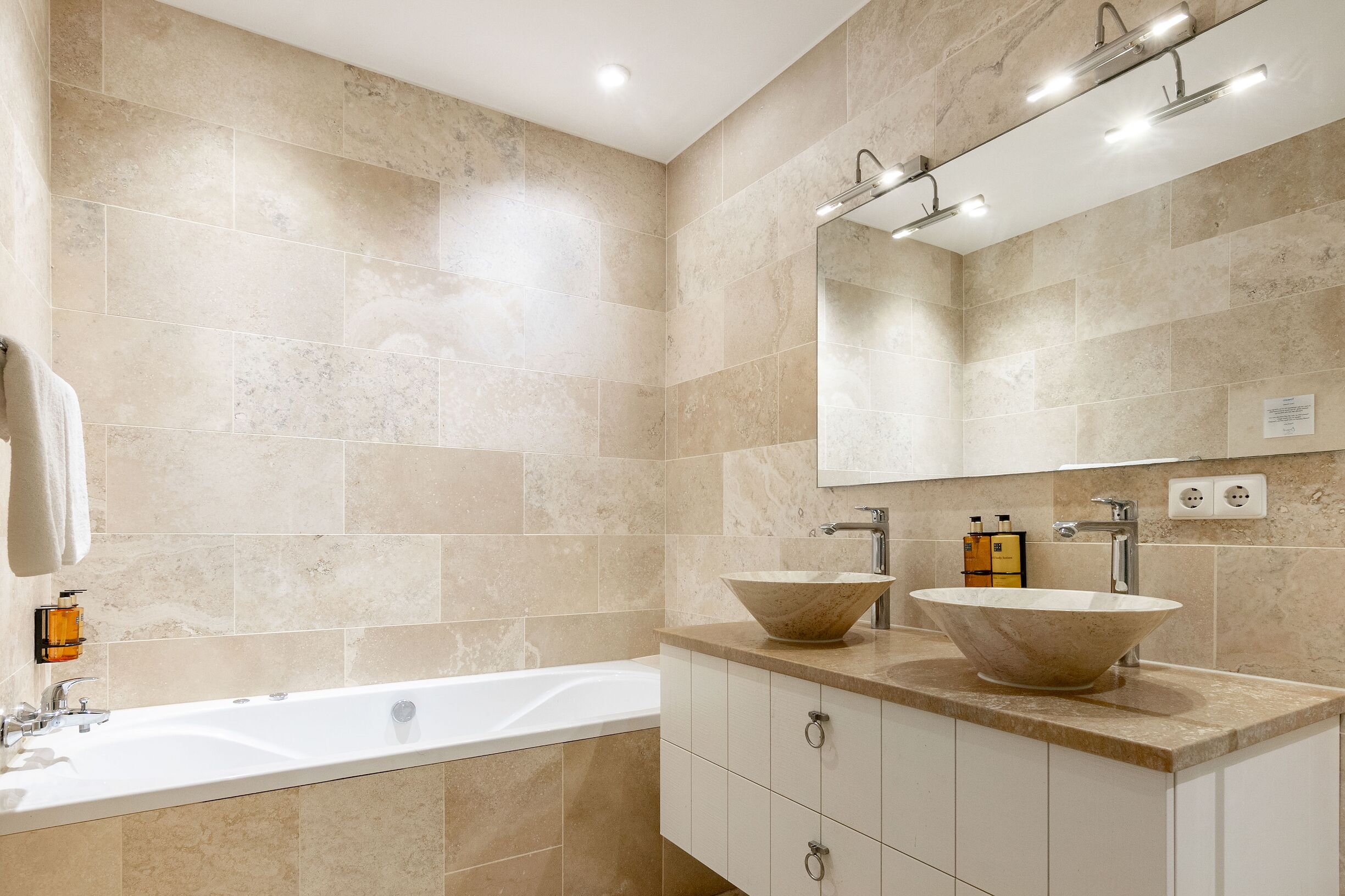 Une salle de bain moderne avec des carreaux de couleur beige, une grande baignoire, deux vasques et un miroir au-dessus du lavabo.