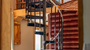 Una escalera en espiral con barandillas de metal que conduce a un nivel superior de madera. Se aprecia una pared de piedra y una puerta de madera al fondo.