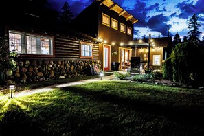 Una cabaña de troncos de dos pisos con cimientos de piedra, un porche delantero y exterior bien iluminado.