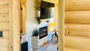 Un interior de madera con chimenea, televisor montado en la pared y una pequeña mesa con lámpara.