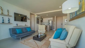 A modern living room with a grey sofa, a white armchair, and a coffee table. There is a flat-screen TV mounted on the wall, and a kitchen area is visible in the background.