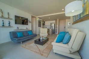 Een moderne woonkamer met een grijze bank, een witte fauteuil en een salontafel. Aan de muur hangt een flatscreen-tv, en er is een keuken te zien op de achtergrond.