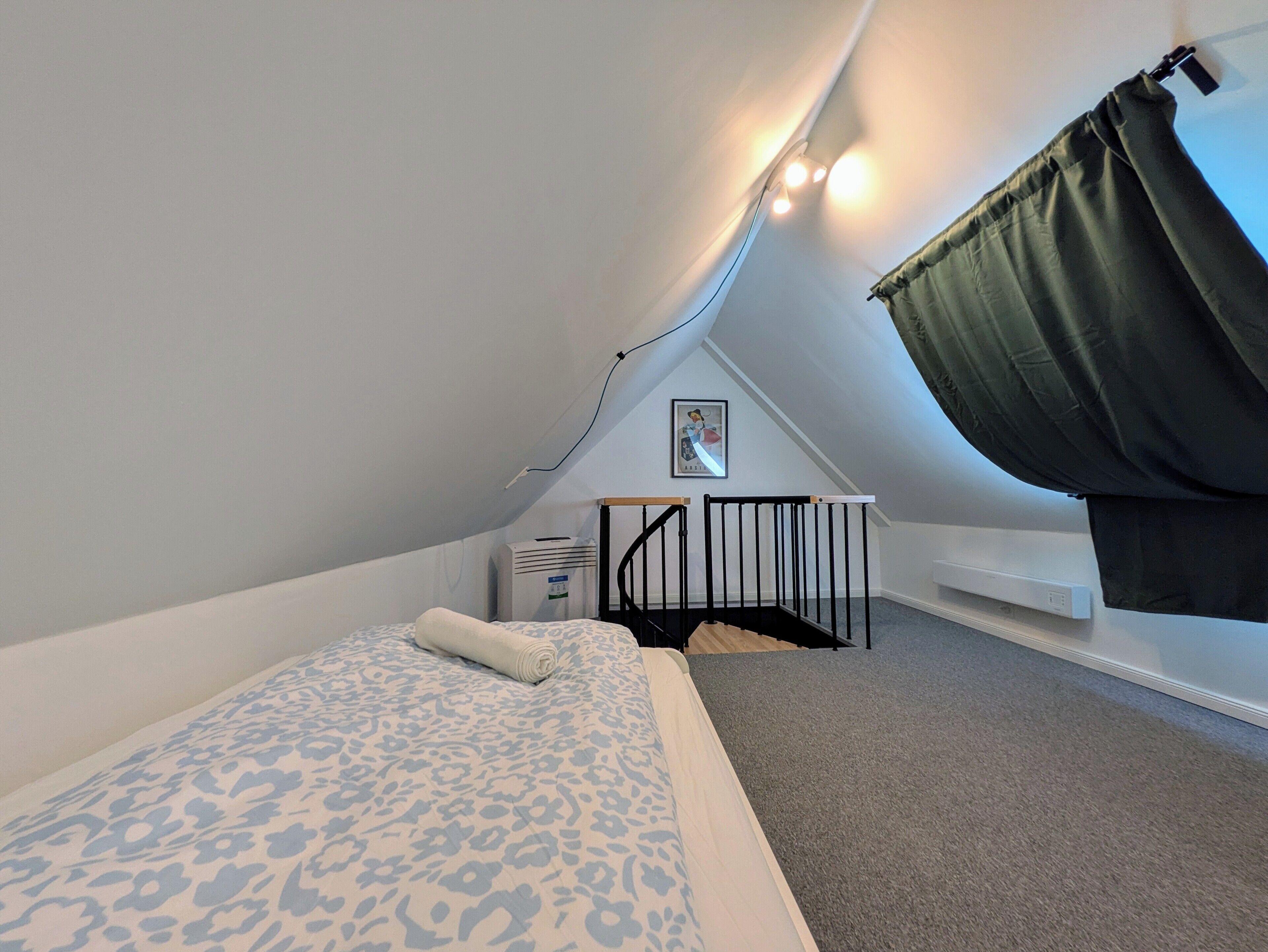 Ein kleines Zimmer mit schrägem Dach, einem Bett, einem Babybett und einem Fenster mit Vorhängen.