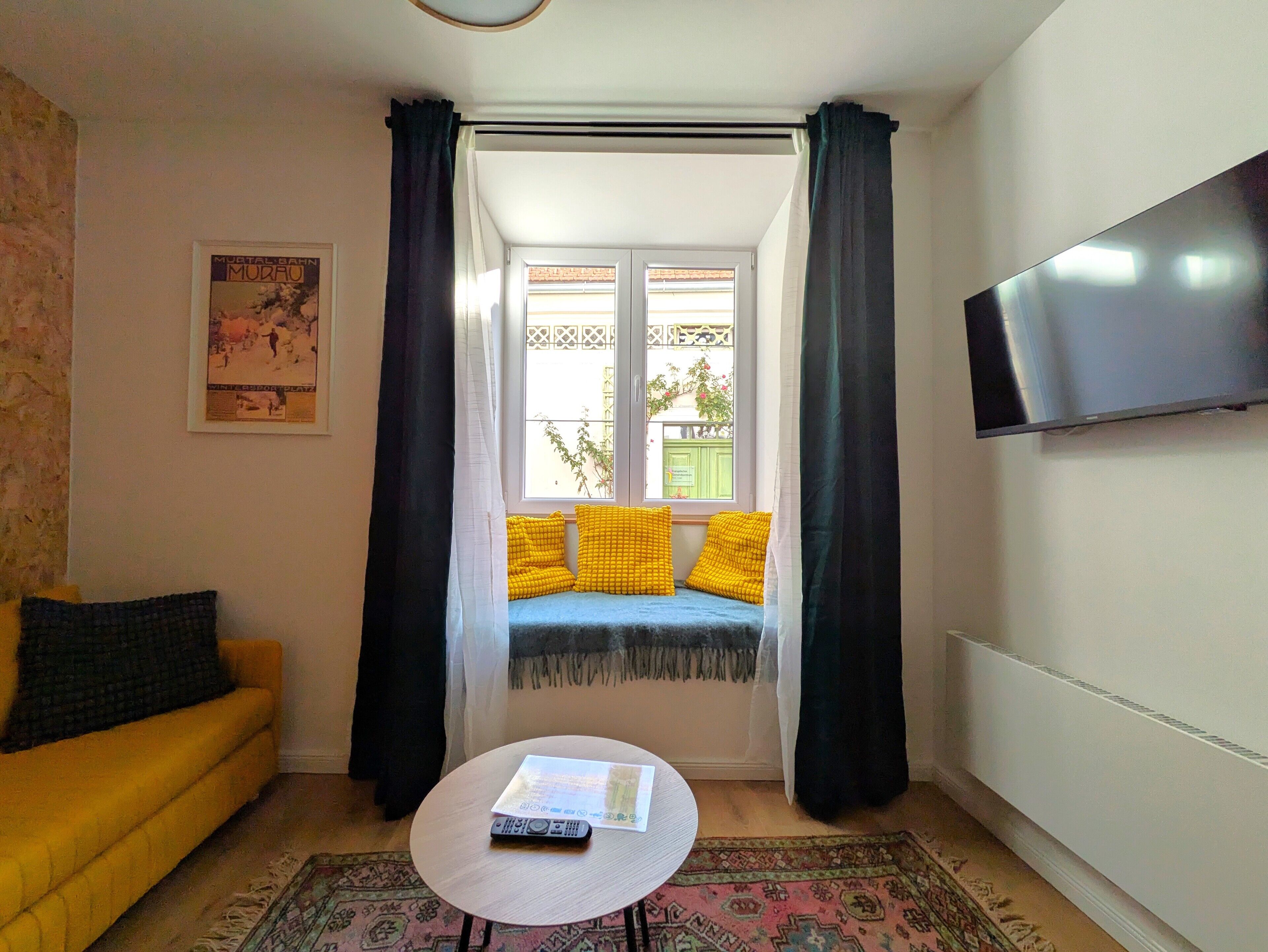 Een kamer met een bank, een kleine tafel met een boek, een aan de muur bevestigde televisie en een raam met gordijnen.