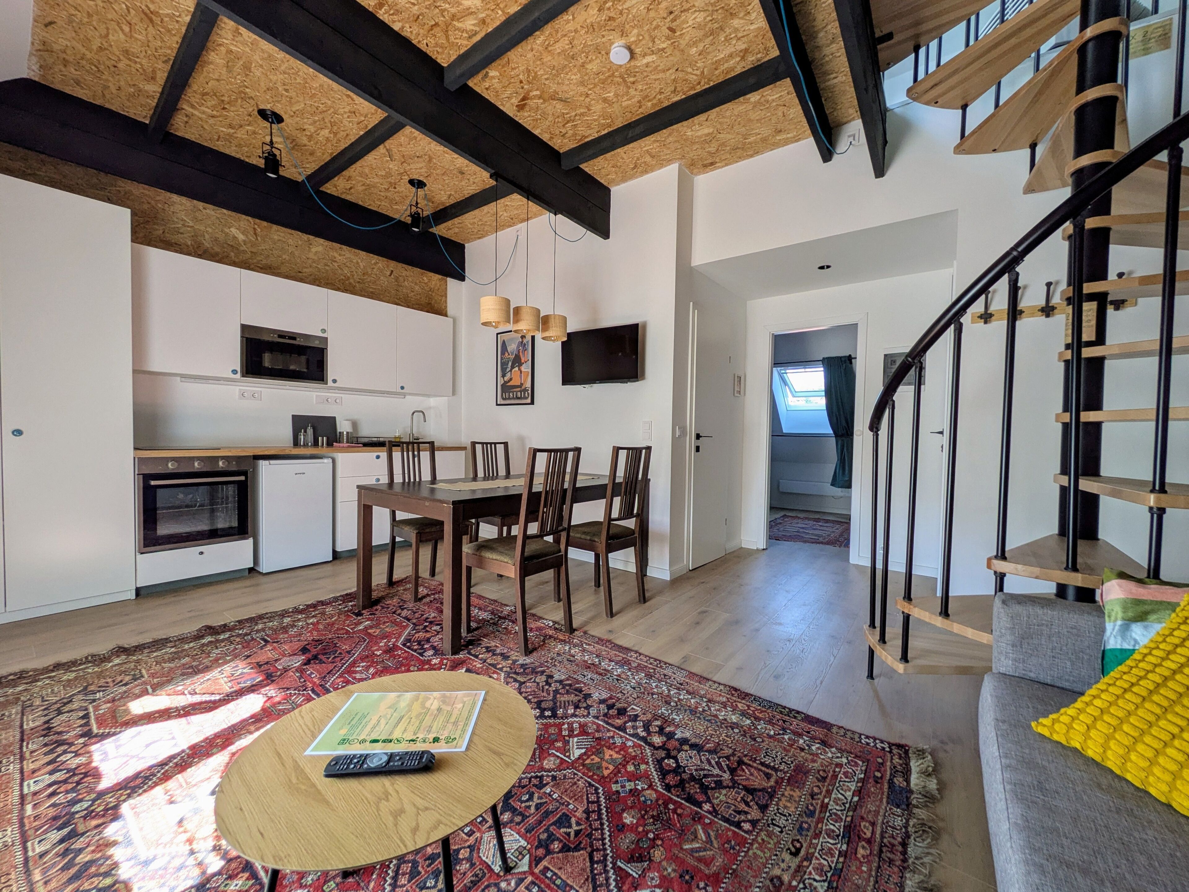 A modern kitchen with white cabinets, a dining area with a wooden table and chairs, a staircase with wooden railings, and a patterned rug on the floor.