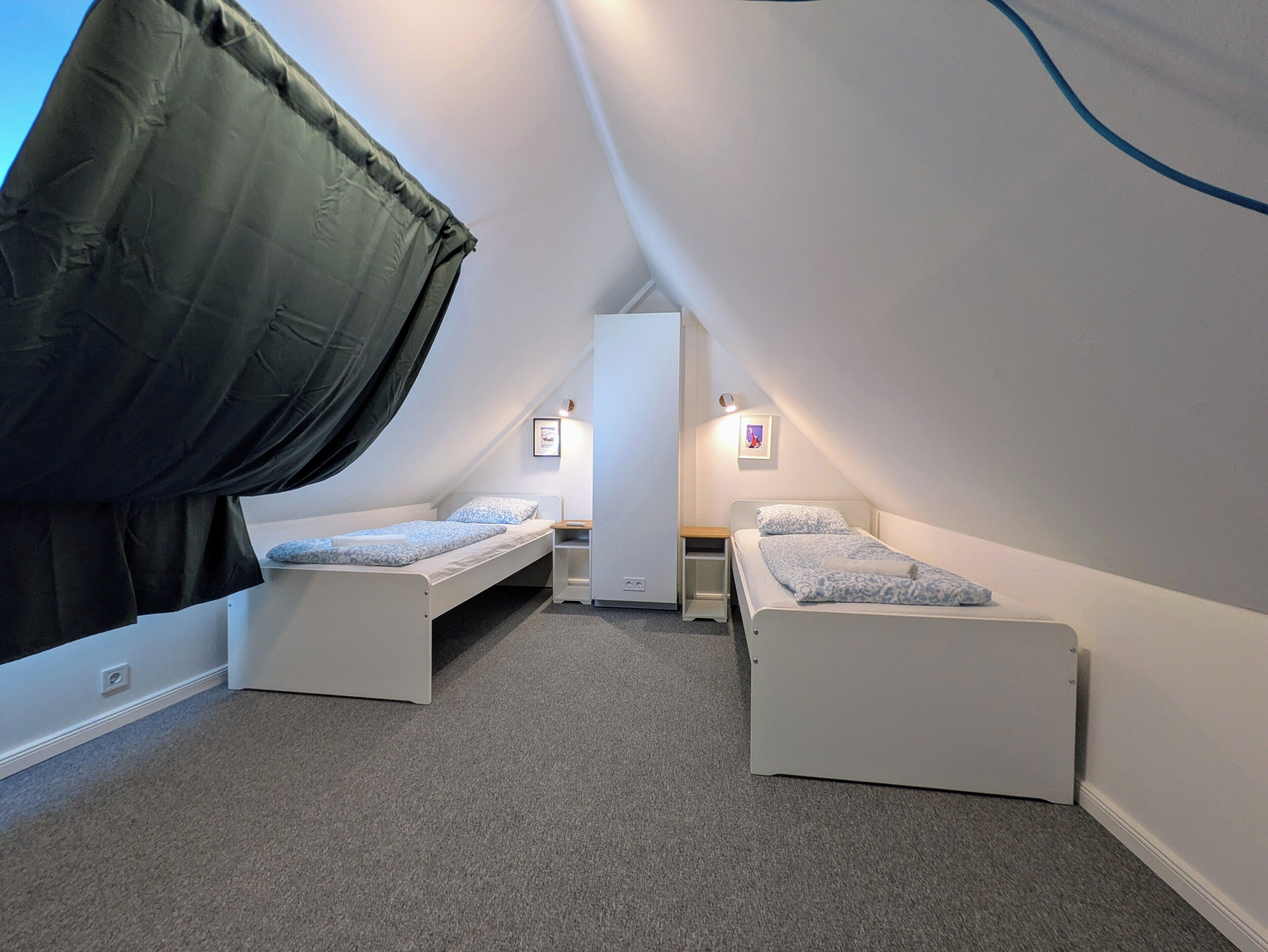 A small, triangular attic bedroom with two single beds, a nightstand, and a wall-mounted light fixture.