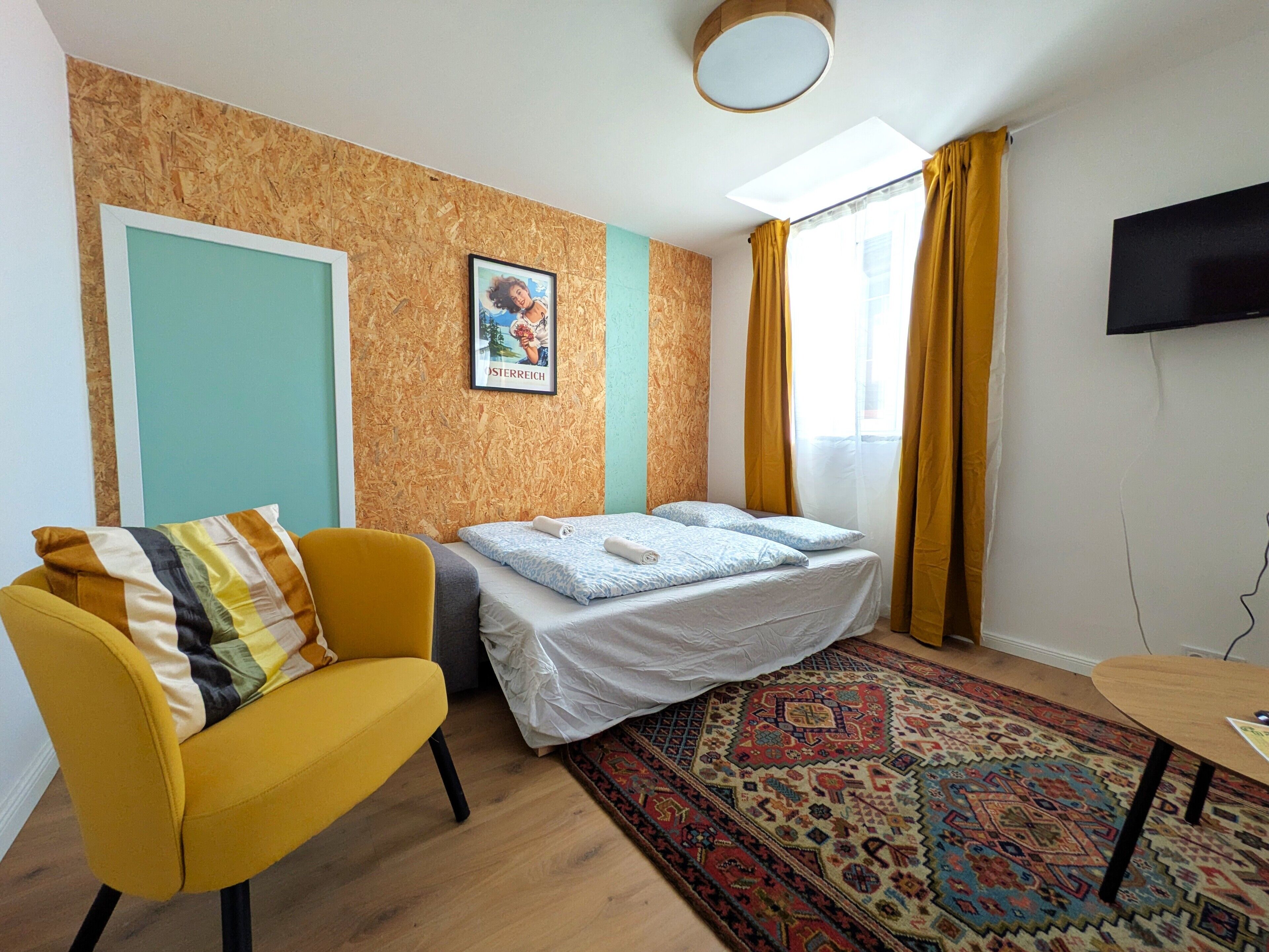 A room with a bed, a yellow armchair, a patterned rug, a wall-mounted picture, and a TV on the wall.