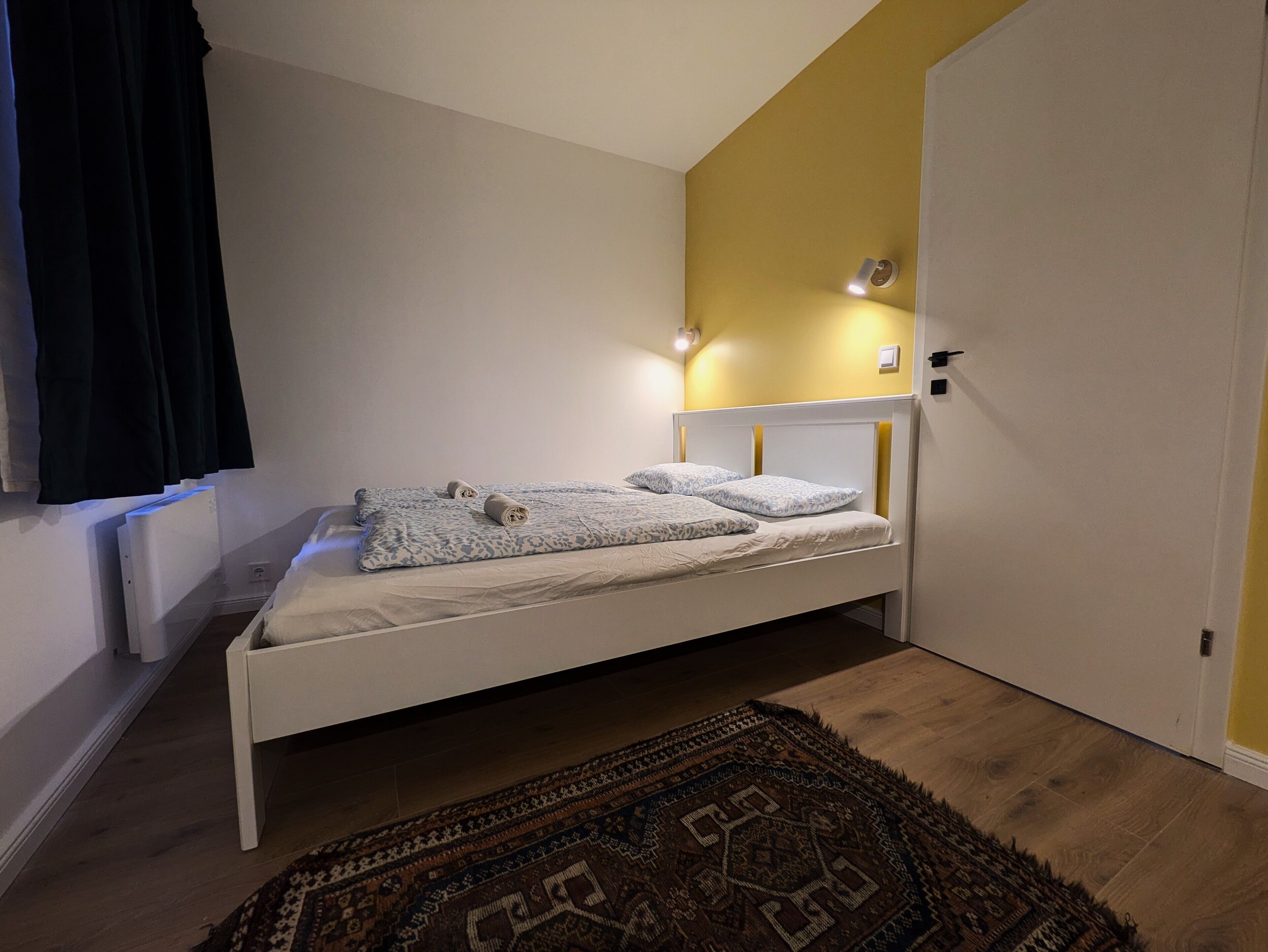 Un lit simple avec une tête de lit blanche et une couvre-lit à motifs, une petite table de chevet et un tapis de couleur sombre sur un plancher en bois.