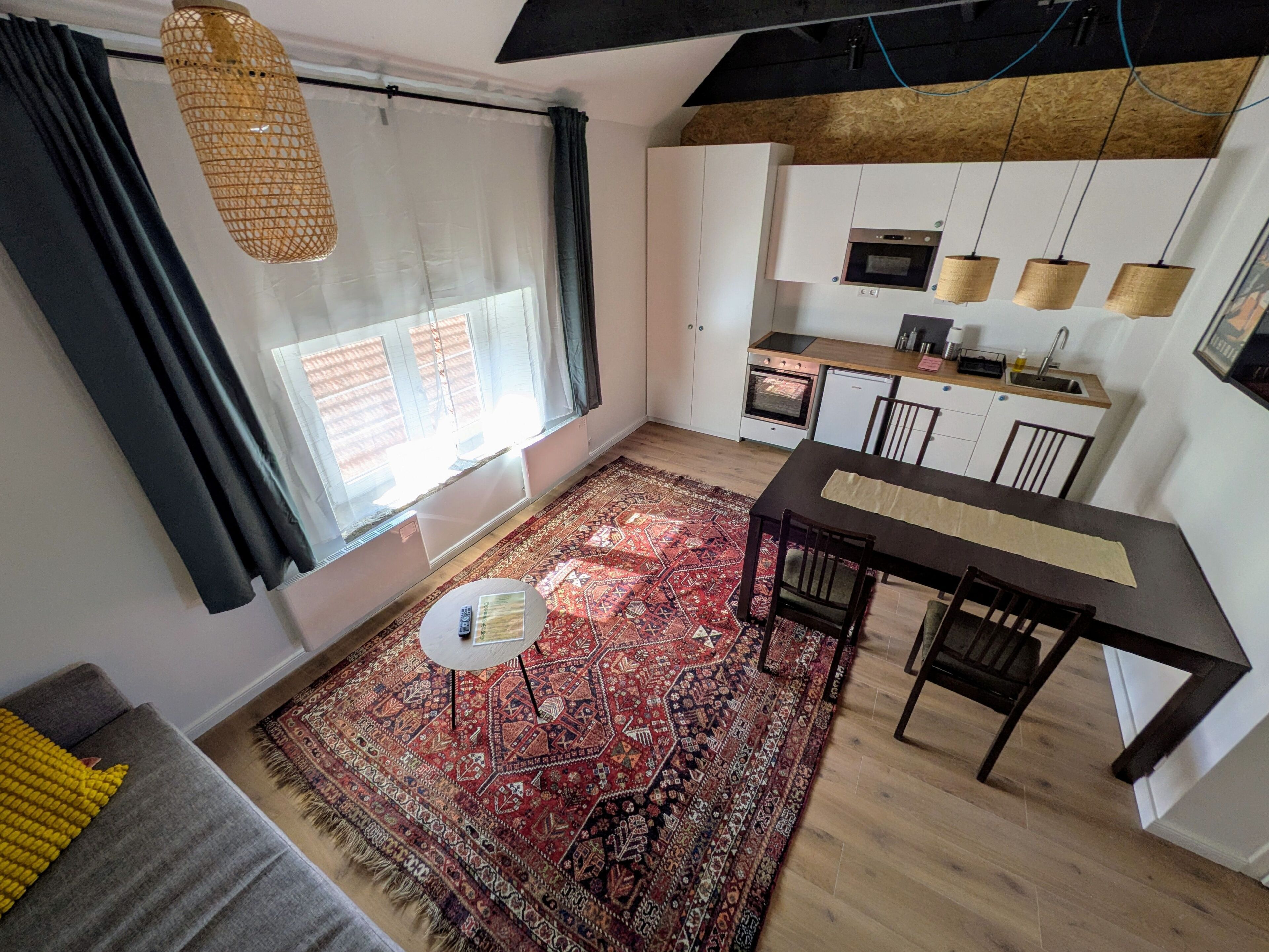 A compact kitchen with a dining area, a large window with curtains, and a patterned rug.