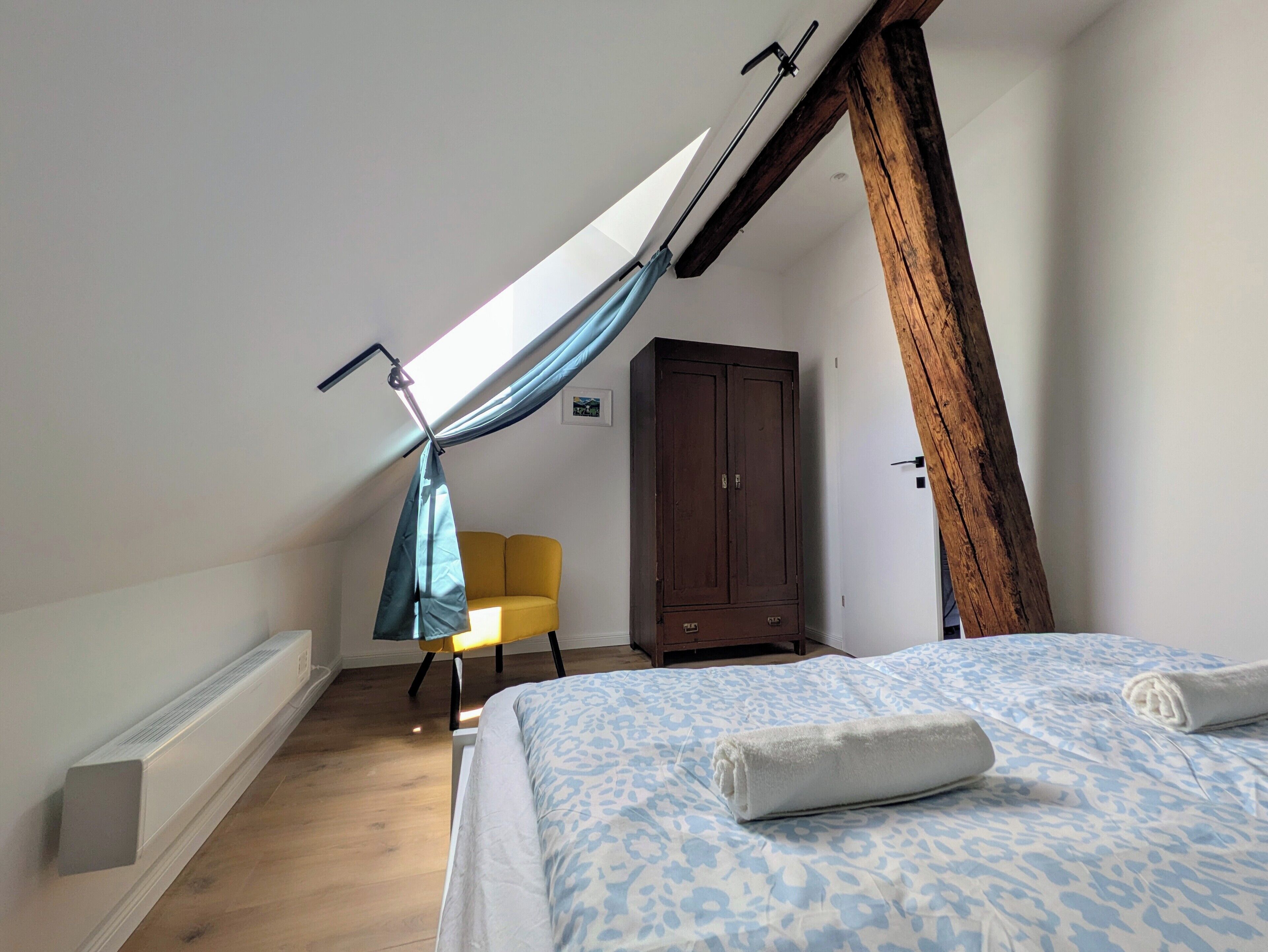 Um quarto com teto inclinado, uma cama com colcha estampada, uma cadeira amarela e um guarda-roupa de madeira.