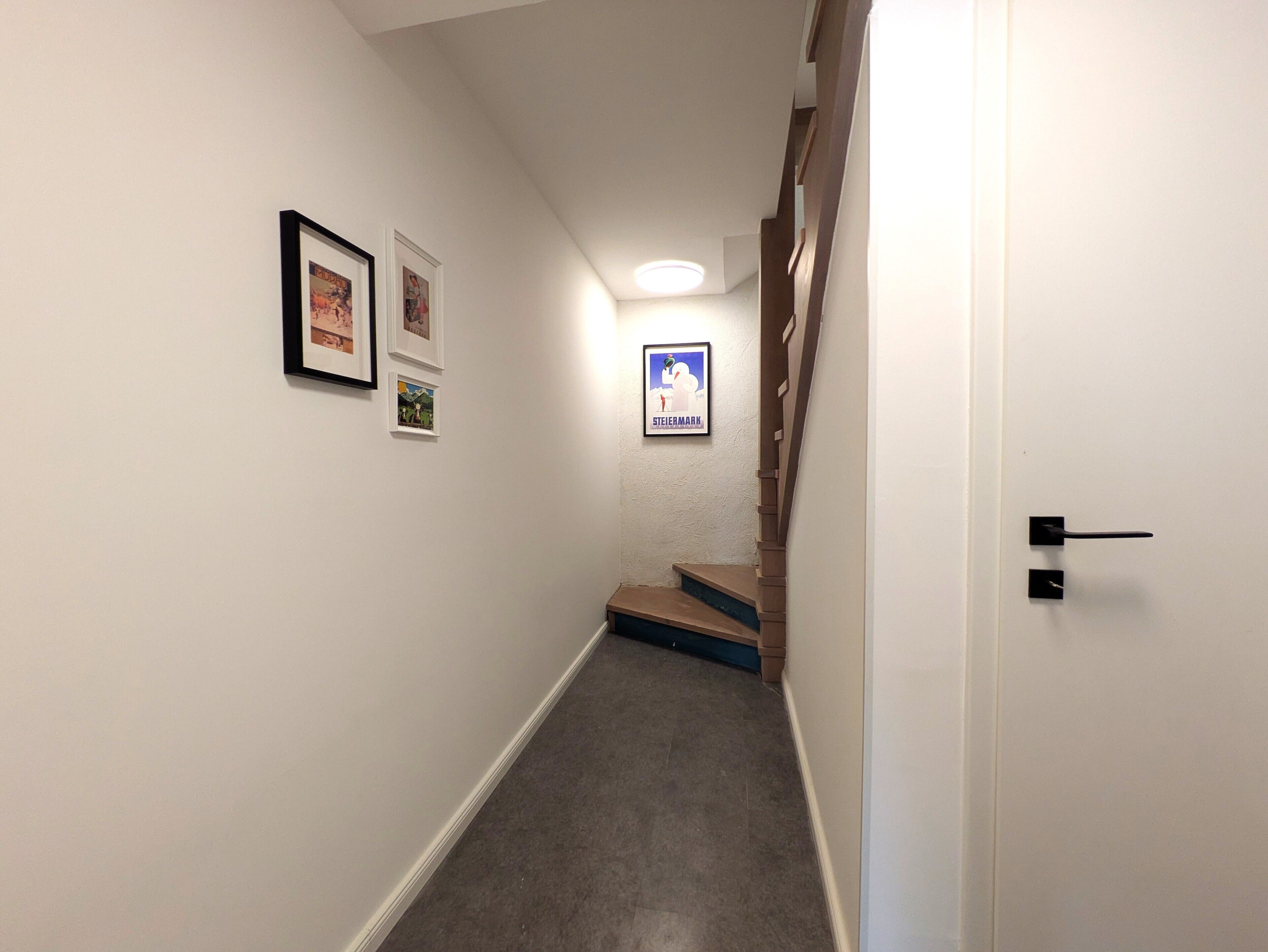 En smal hall med vita väggar, ett mattbeklätt golv och en trätrappa som leder upp till en liten plattform. På väggarna hänger inramade tavlor och det finns en dörr med ett svart handtag.