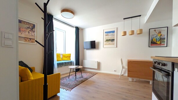 A modern apartment with a living area featuring a yellow sofa, a dining table, and a kitchen with a stove. - mg4 Apartments (Murau)